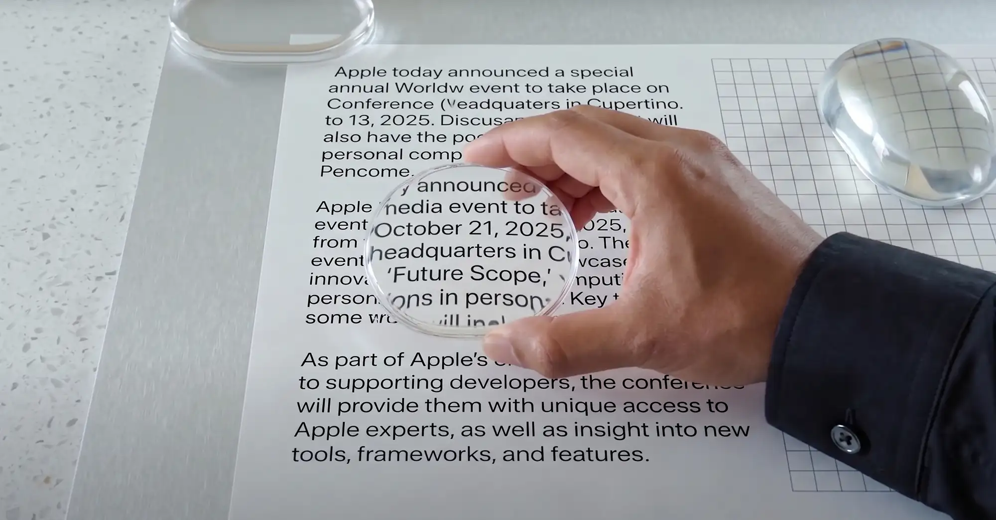 a close-up, high-angle studio photograph of a person's hand in a black shirt holding a round magnifying glass over a piece of white paper with black text, also visible is a transparent, grid-patterned mouse and another magnifying object, all dramatically lit on a light gray speckled surface