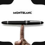 product photography of a montblanc fountain pen, polished black with silver accents, horizontally balanced on a single finger against a clean white background, black montblanc logo above, with stylized black and white textured montblanc star emblem framing the shot, soft, even studio lighting